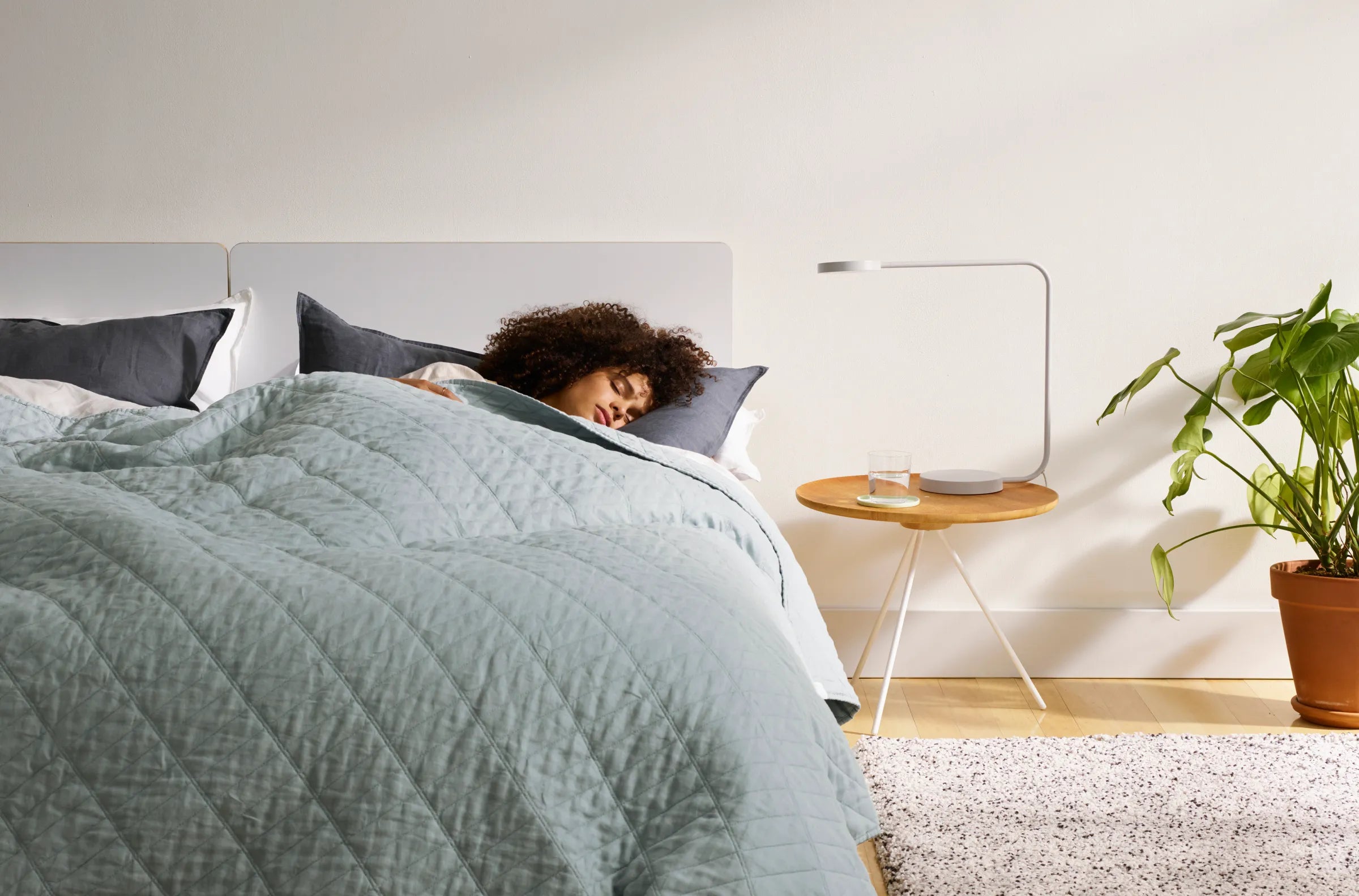 Person lying in bed under a light green comforter with a plant and side table in the background.